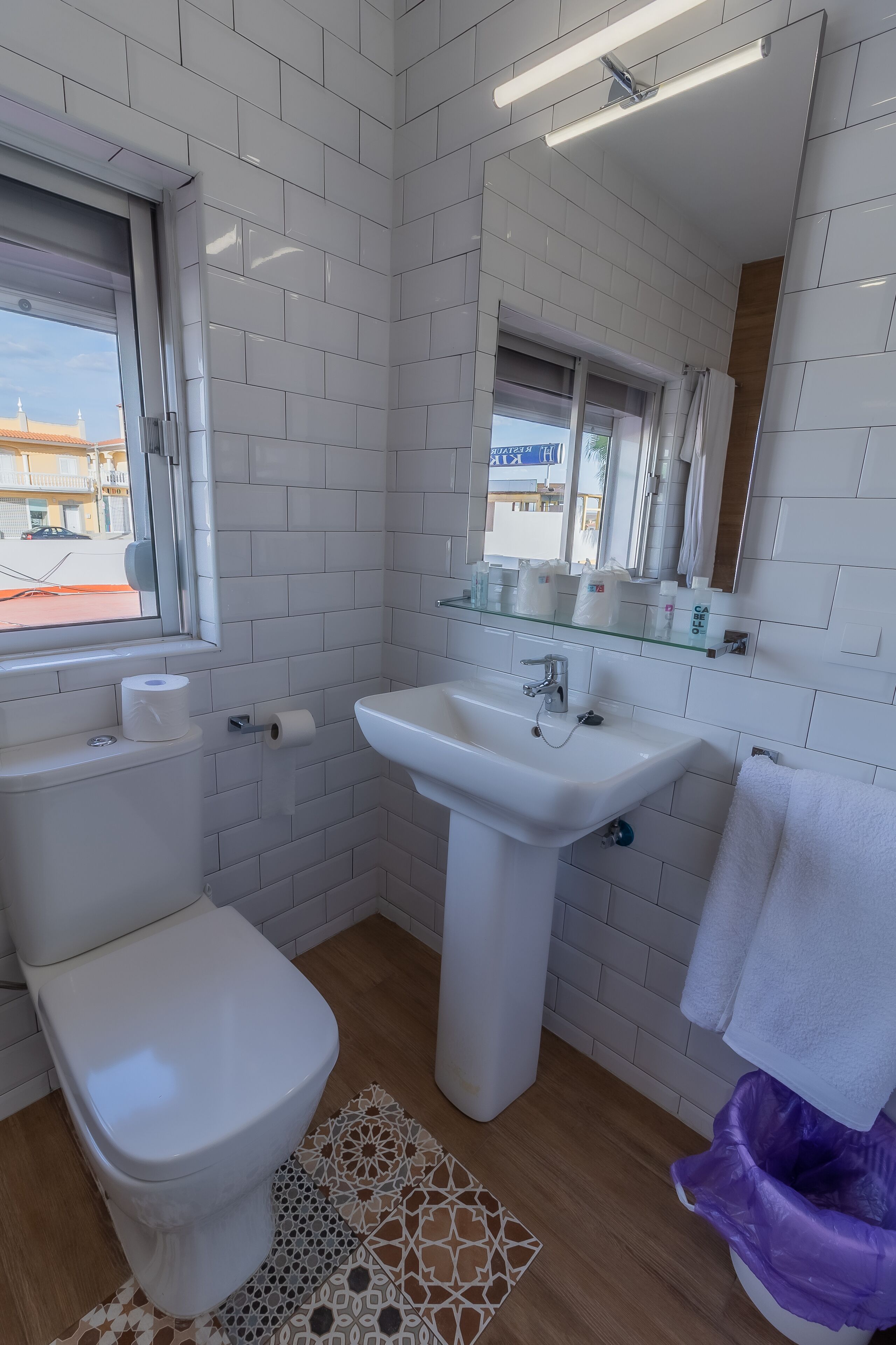 Un baño con baldosas blancas, un lavamanos blanco, un inodoro, una ventana y un espejo.