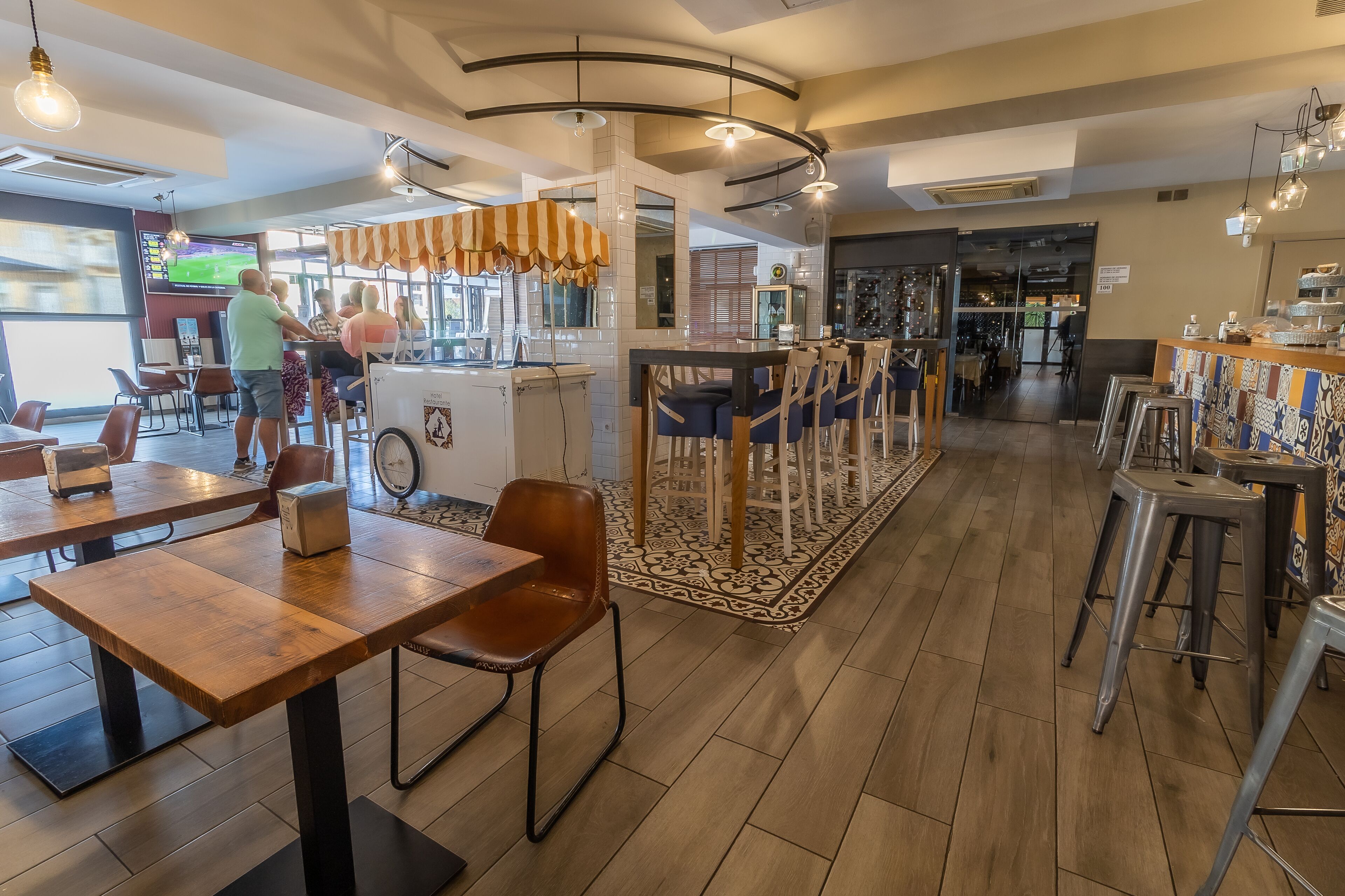 Un café moderno con mesas y sillas de madera, una barra con un paraguas a rayas y una gran pantalla de televisión mostrando deportes.