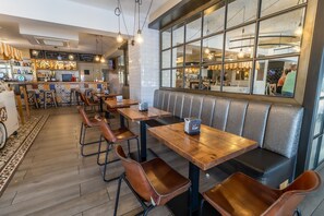 Un interior de restaurante con mesas de madera y asientos de cuero, una zona de bar con botellas y un amplio ventanal con vistas a la calle.