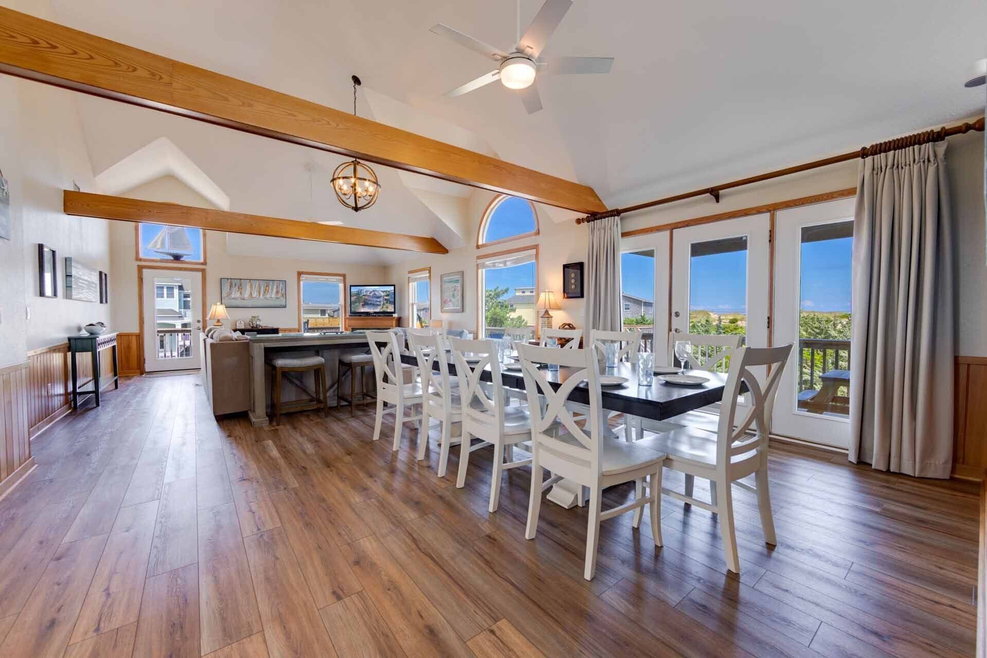 A spacious kitchen with wooden beams, a dining area with white chairs, and large windows offering a view of the landscape.