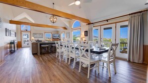 Une cuisine spacieuse avec des poutres en bois, un coin repas avec des chaises blanches et de grandes fenêtres offrant une vue sur le paysage.