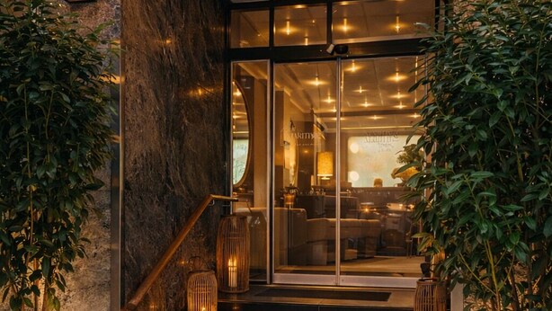 Entrada de hotel con una escalera que conduce a una recepción, plantas en macetas e iluminación en la pared.