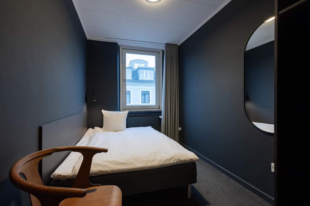 A hotel room with a bed, a chair, a window with curtains, and a round mirror on the wall.