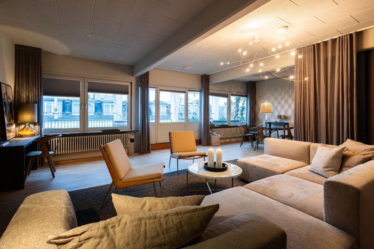 A modern living room with a sofa set, a dining area, and large windows offering a view of the city.