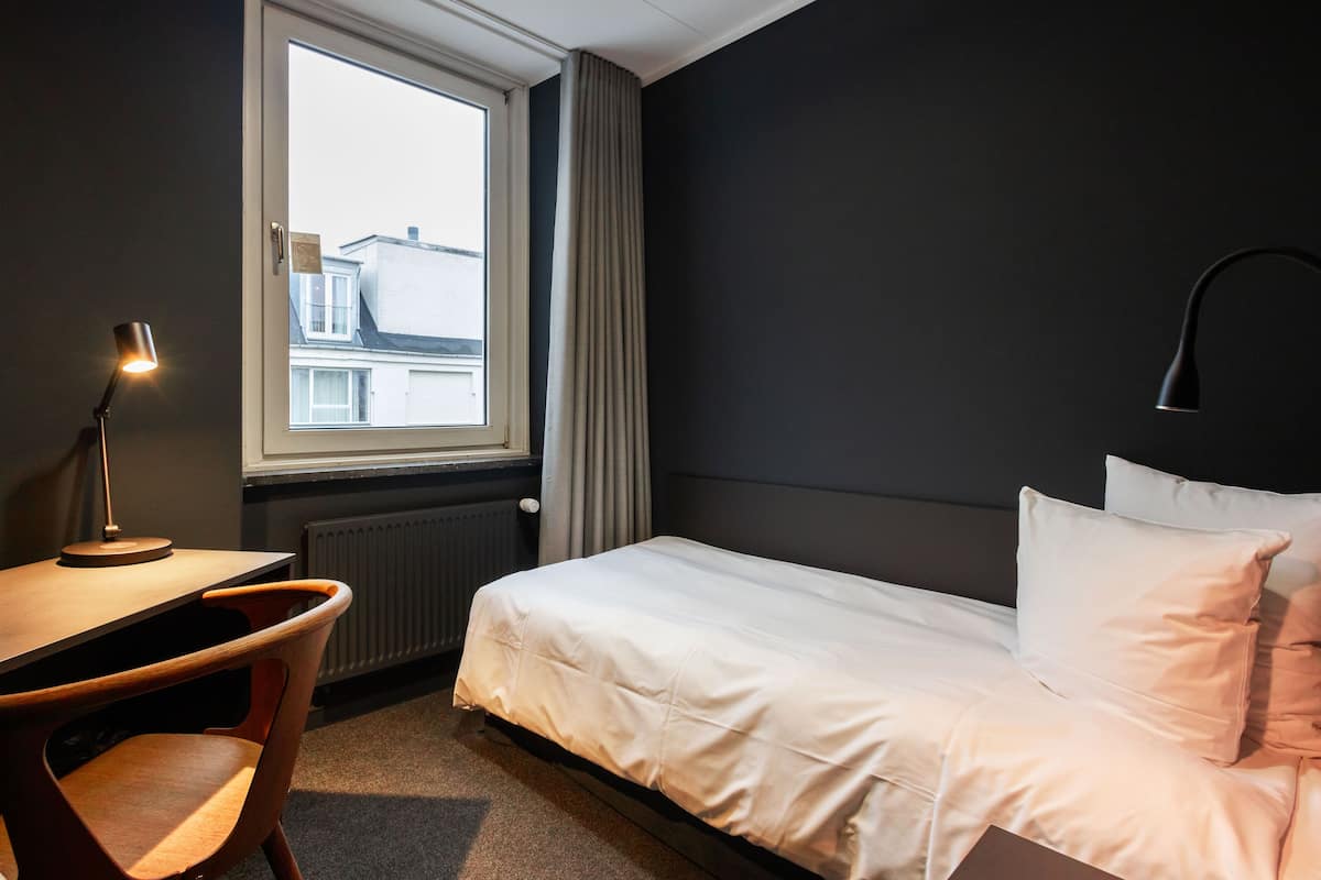 A hotel room with a bed, a desk, a chair, a window, and a radiator.