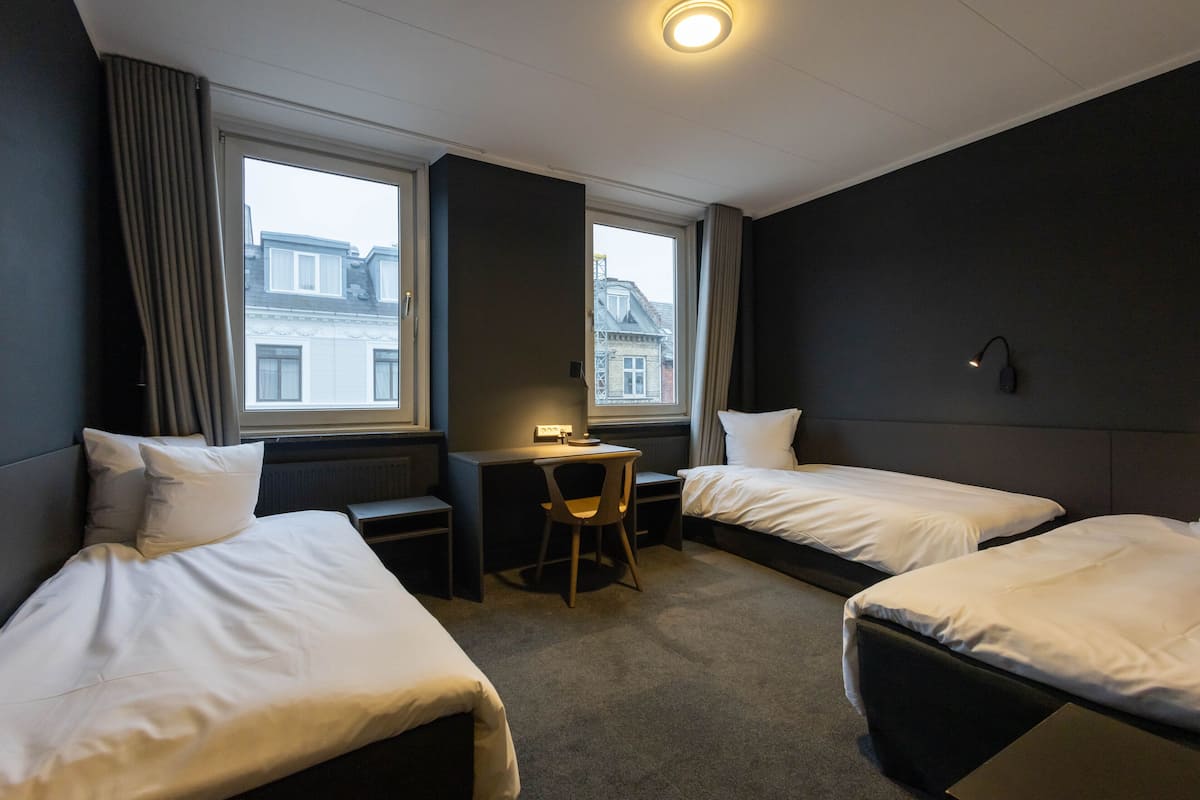 A hotel room with two single beds, a desk with a chair, and a large window providing a view of buildings outside.