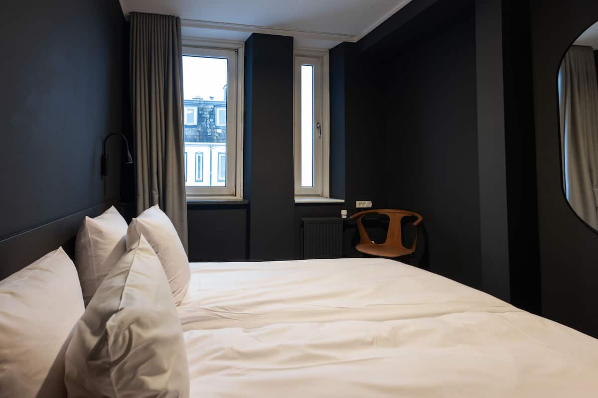 A hotel room with a large bed, a chair, a window with curtains, and a view of buildings outside.