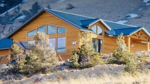 A wooden cabin with a sloped roof, large windows, and a deck area. - Experience the Magic of Wapiti Valley in Luxury. Stunning Views-East Yellowstone (Cody)
