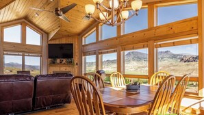 A cozy cabin interior with a wooden ceiling, large windows offering a view of a mountainous landscape, a dining table with chairs, and a leather sofa. - Experience the Magic of Wapiti Valley in Luxury. Stunning Views-East Yellowstone (Cody)