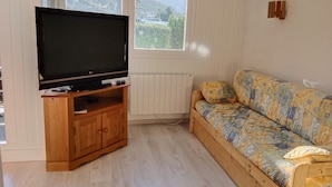 Una habitación con televisión sobre un mueble de madera, un sofá con cojines estampados y una ventana que ofrece vistas a zonas verdes y montañas.