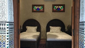 Quarto com duas camas, cada uma com cabeceira e travesseiro, e um chão com padrão. Há duas janelas pequenas com painéis de vidro decorativo na parede.