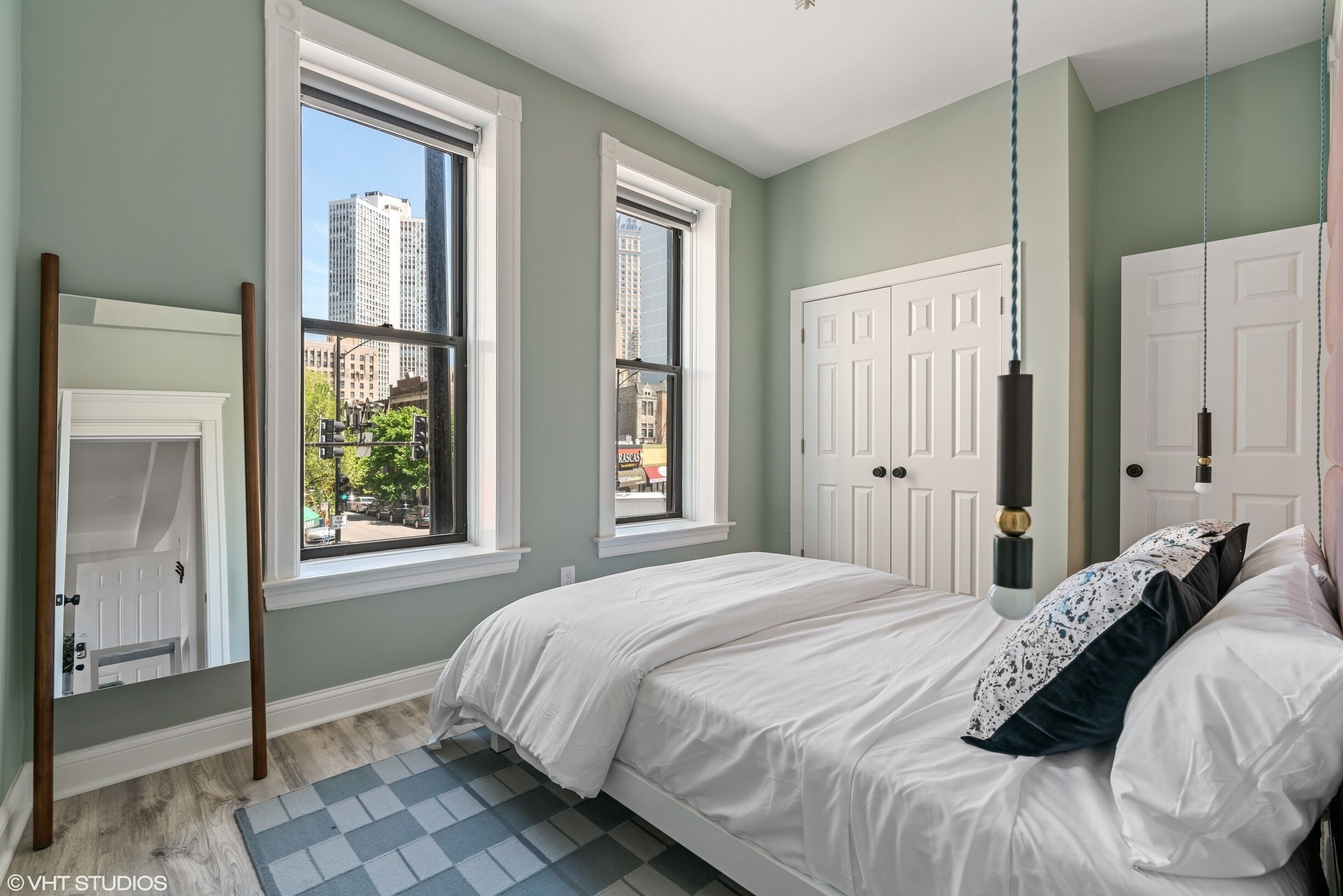 A bedroom with a bed, two windows, a door, and a view of buildings outside.