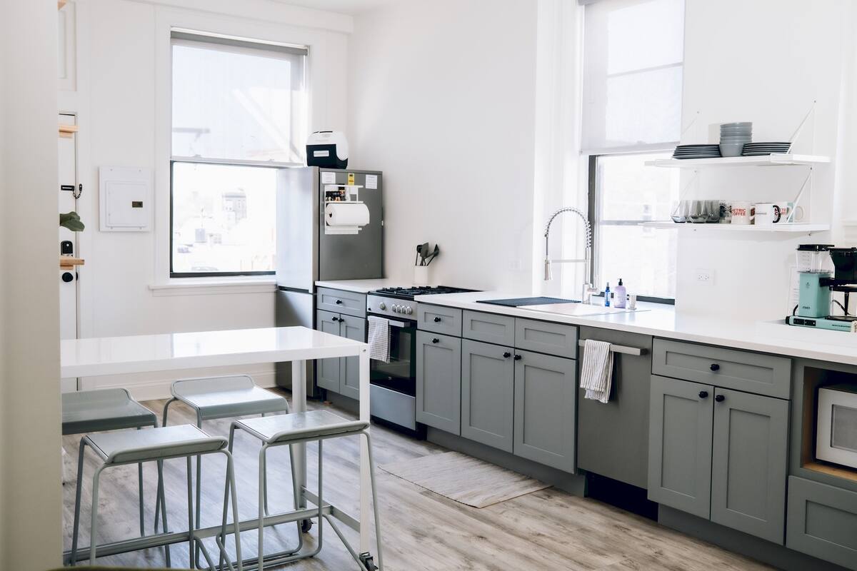 Une cuisine moderne avec des meubles gris, un plan de travail blanc et un four encastré. On y trouve des tabourets de bar et une fenêtre qui laisse entrer la lumière naturelle.