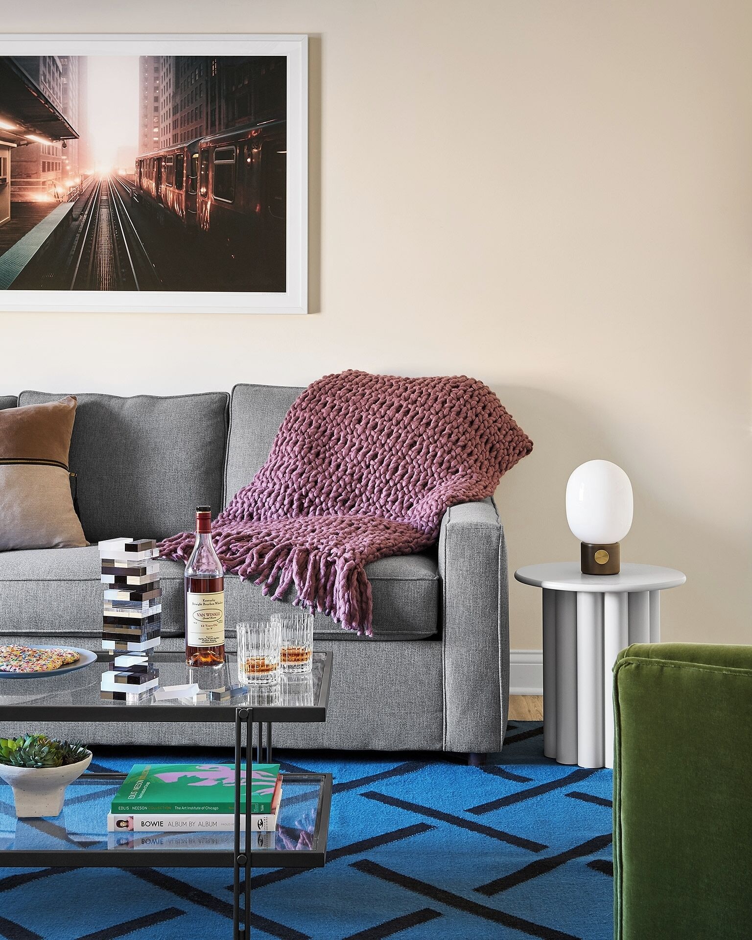 A modern living room with a grey sofa, a coffee table with books and a bottle, a green armchair, a round side table with a lamp, and a framed cityscape painting on the wall.