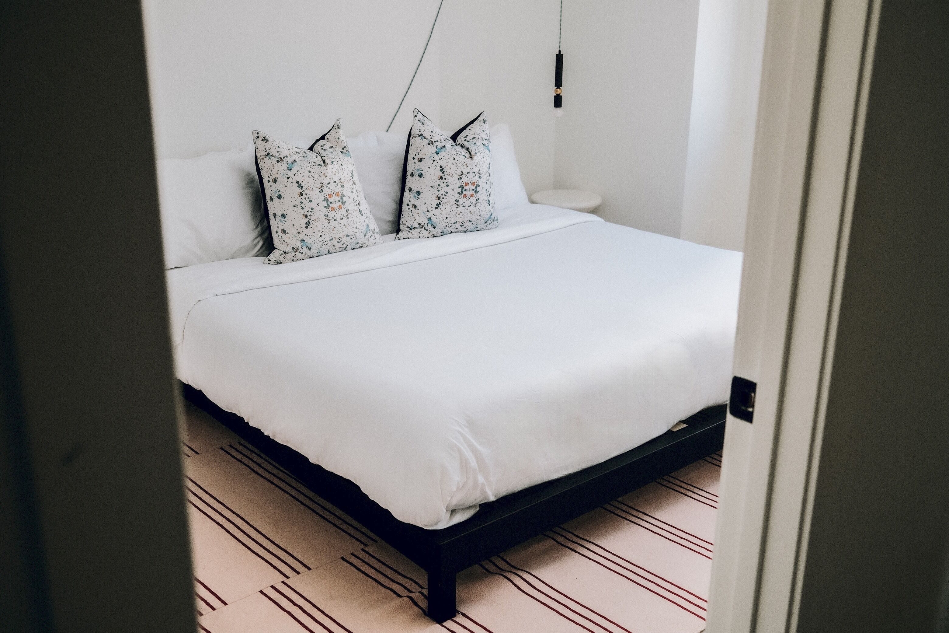 Una cama bien hecha con ropa blanca y almohadas estampadas con motivos florales, una mesita de noche con una lámpara y un suelo a rayas.