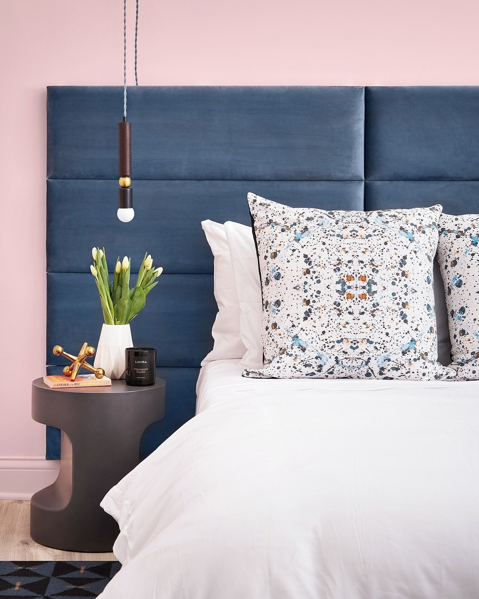 Een slaapkamer met een blauw bekleed hoofdeinde, wit beddengoed, een gestreept kussen, een nachtkastje met een bloevas en een lamp.