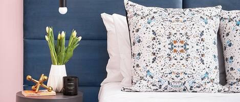 Een slaapkamer met een blauw bekleed hoofdeinde, wit beddengoed, een gestreept kussen, een nachtkastje met een bloevas en een lamp.
