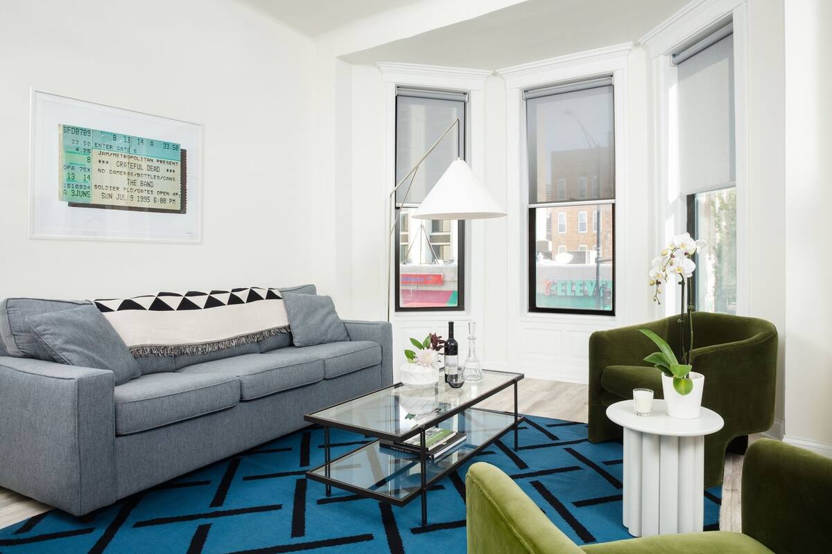 Un salon moderne avec un canapé gris, une table basse en verre et un fauteuil vert. Un billet encadré est accroché au mur, et une fenêtre offre une vue sur les bâtiments.