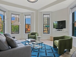 Una sala de estar moderna con un sofá, dos sillones, una mesa de centro, un televisor y ventanas amplias con vistas a la ciudad.
