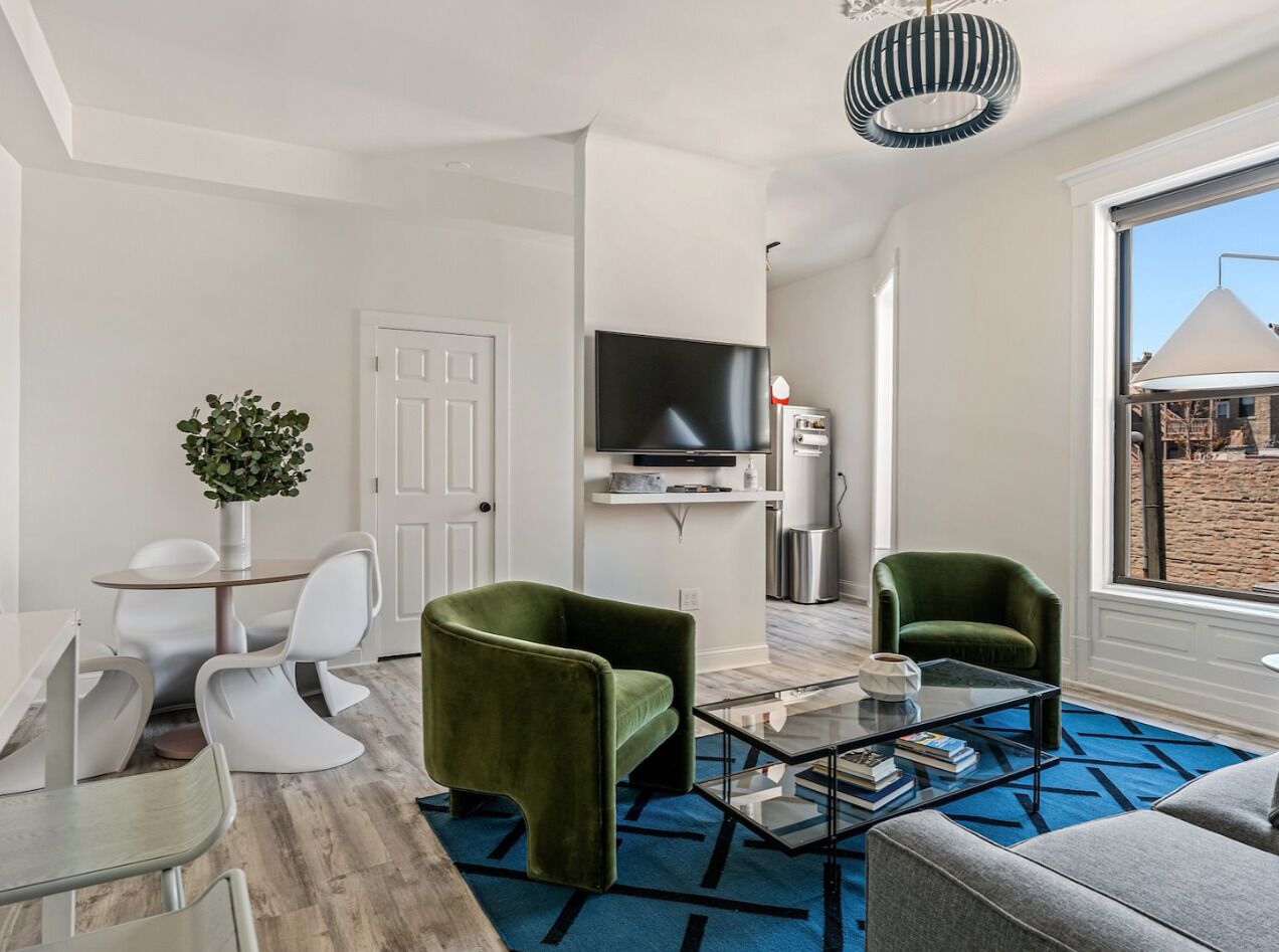 Un salon moderne avec un téléviseur, des fauteuils verts, une table basse en verre et une vue sur l’extérieur.