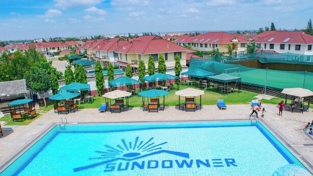 Ein Schwimmbecken mit der Aufschrift "SUNDOWNER" und einem Sonnendesign, umgeben von Liegestühlen und Sonnenschirmen, mit Wohnhäusern im Hintergrund.