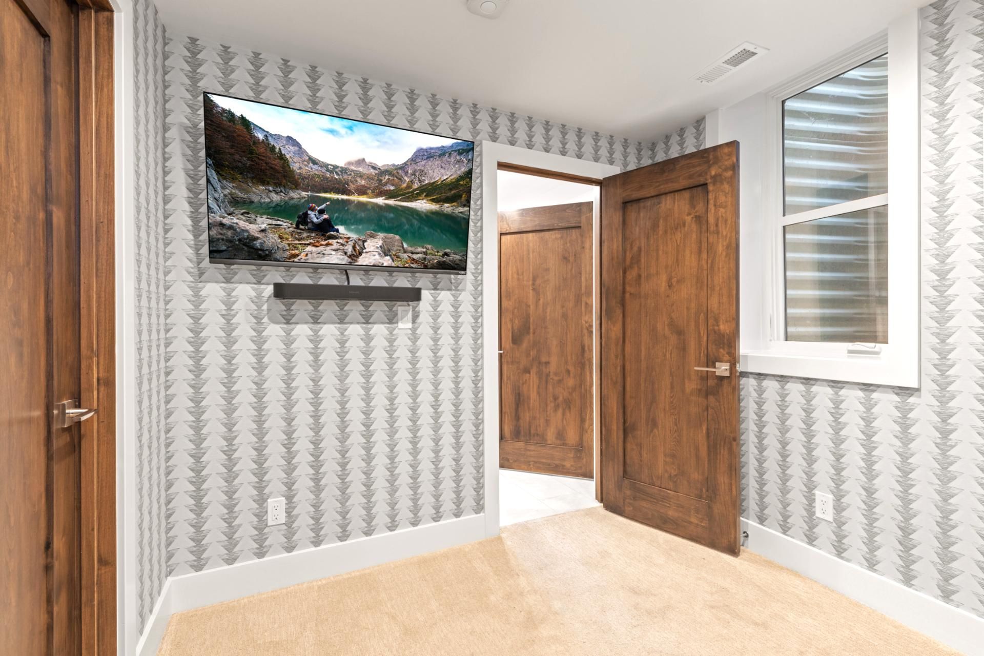 Un couloir avec un papier peint à motifs, un téléviseur fixé au mur diffusant un paysage, une porte en bois et une fenêtre avec des stores.