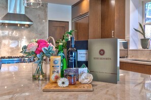 A kitchen counter with a variety of items including a bottle of olive oil, a box of crisps, a bottle of water, a bottle of wine, and a decorative flower arrangement. - Walk to Ski Slopes + Trails! Luxury 4-Bedroom Private Home with Private Hot tub, A/C, Heated Parking, and Two Master Suite! | PEA-1009 by KBM Resorts (Park City)
