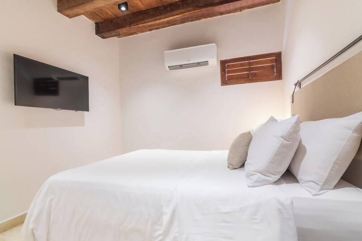 Une chambre à coucher avec un lit, du linge de lit blanc, un téléviseur fixé au mur, un appareil de climatisation et une poutre en bois au plafond.