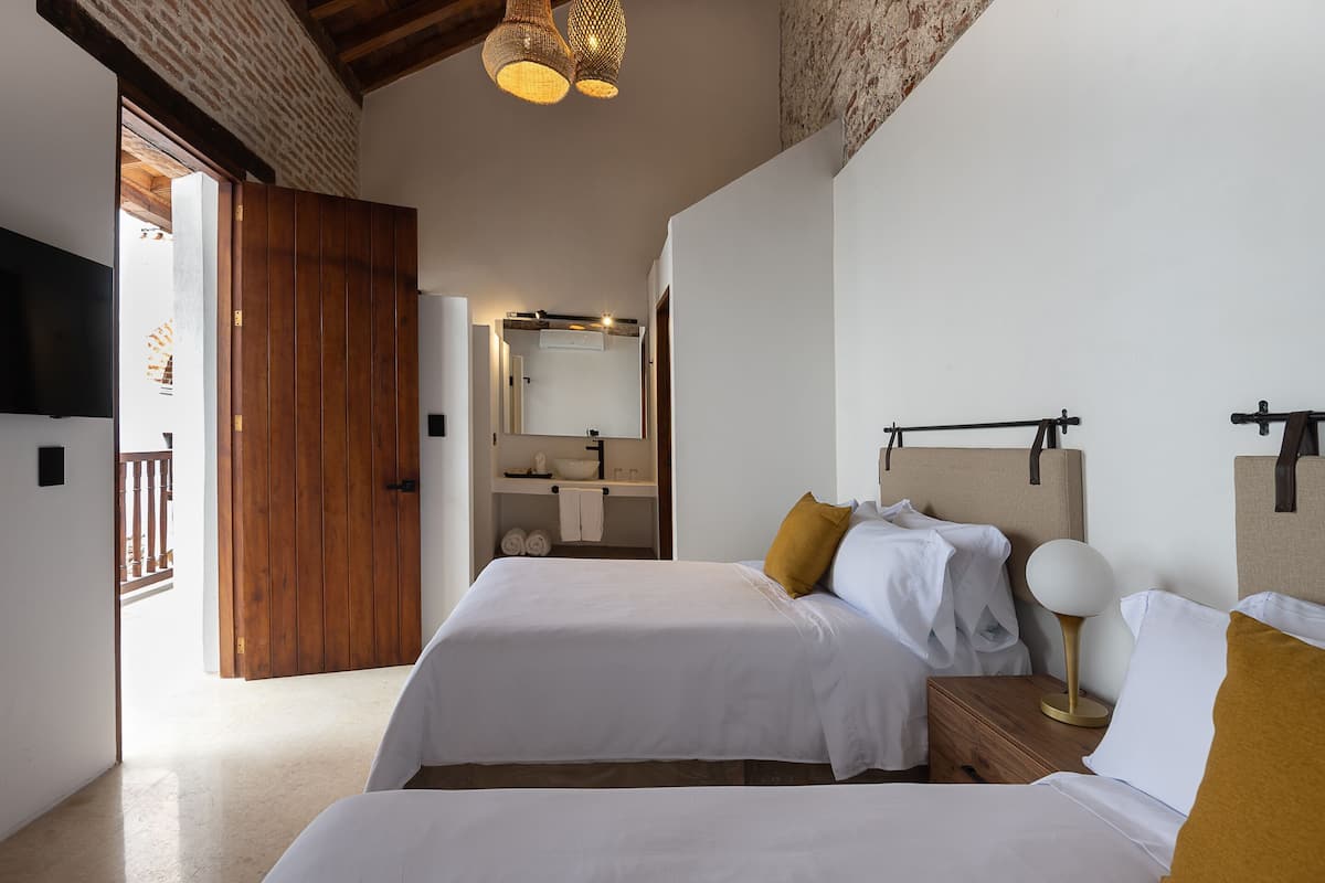 Une chambre avec deux lits, une armoire en bois, une table de chevet avec une lampe, et une salle de bain visible à travers une porte ouverte.