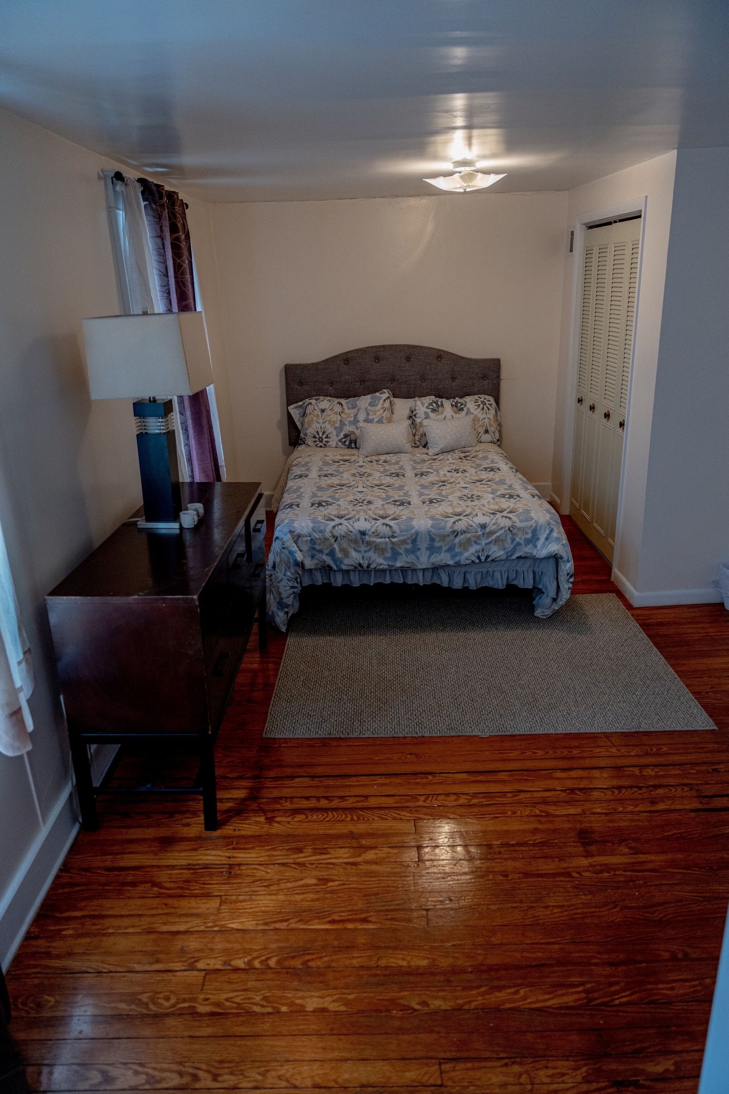 Een slaapkamer met houten vloer, een bed met een bloemenprint beddegoed, een nachtkastje met een lamp en een garderobe met verstelbare deuren.