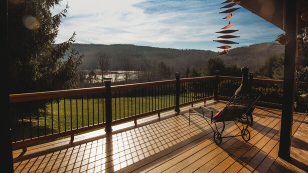 A wooden deck with a view of a green field and trees, a baby stroller, and a barbecue grill.
