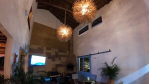 A spacious indoor area with a high ceiling, rustic wooden beams, and hanging light fixtures. There is a television mounted on the wall, a dining table with chairs, and several potted plants.