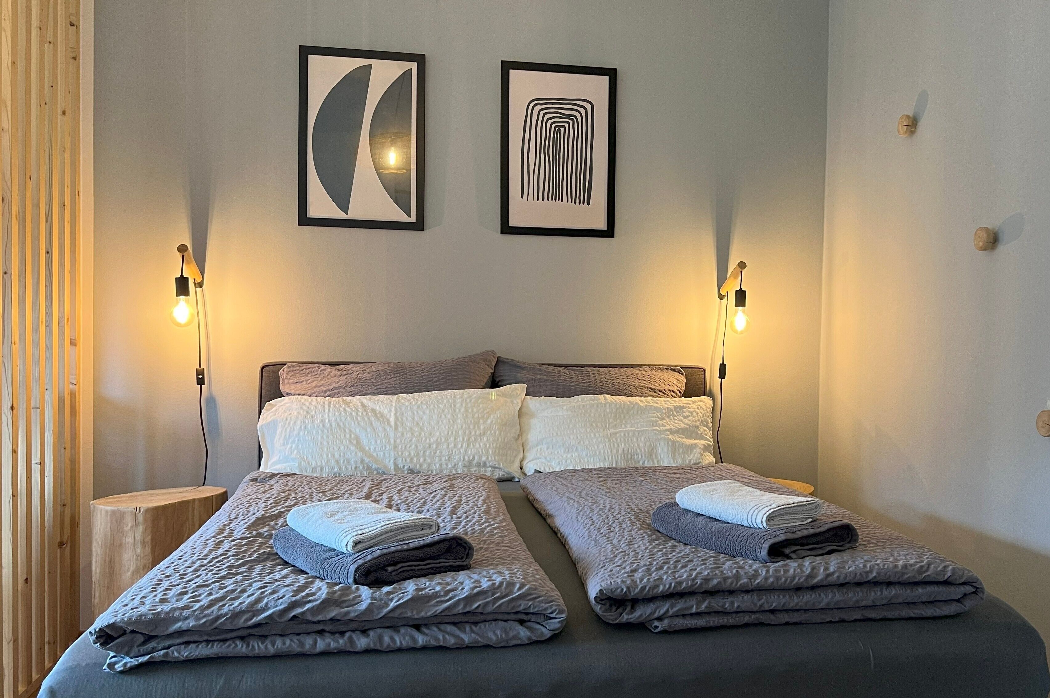 Una cama doble con dos almohadas, una mesita de noche, dos lámparas de pared y dos obras de arte enmarcadas.