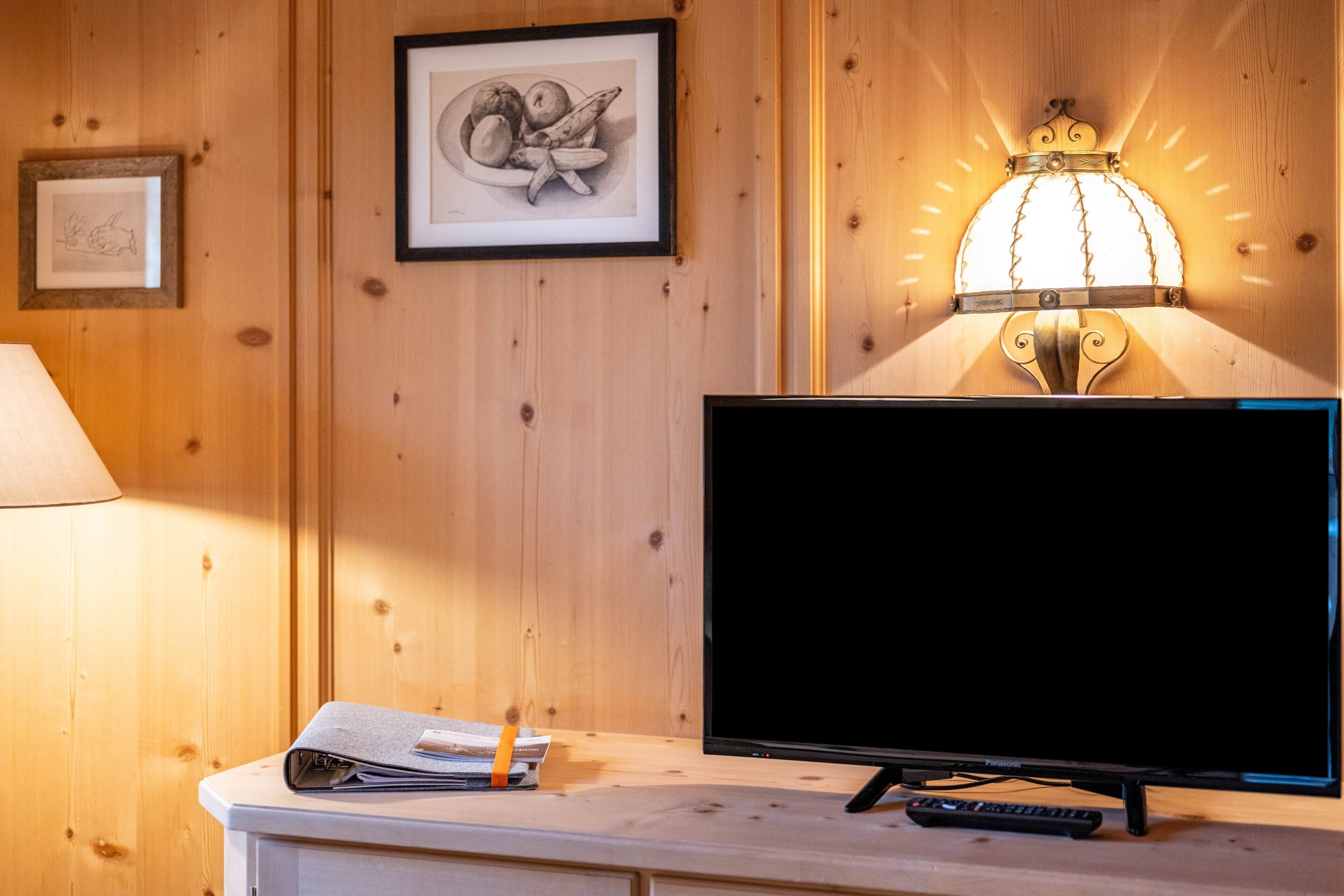 Un mur en bois orné de tableaux encadrés, une applique murale éclairée, un téléviseur et une télécommande posés sur une surface blanche.
