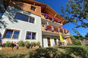 Ein zweigeschossiges Gebäude mit großen Fenstern, einem Balkon mit Blumenkästen und einem grünen Sonnenschirm.