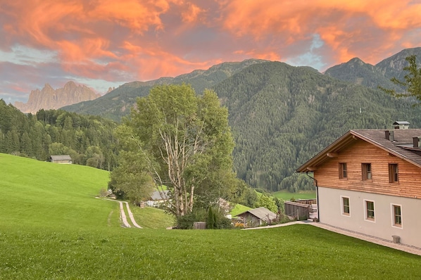 A rural house with a wooden exterior, surrounded by green fields and mountains.