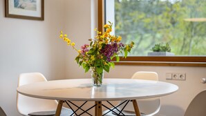 Ett runt bord med en vas med blommor, två stolar, en hängande lampa och ett fönster med utsikt över grönområden.