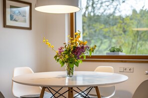 A round table with a vase of flowers, two chairs, a hanging light fixture, and a window with a view of greenery.