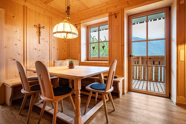 Ein Holzinnenraum mit einem Esstisch, Stühlen, einer Deckenleuchte, einem Kruzifix an der Wand und einem großen Fenster mit Ausblick.