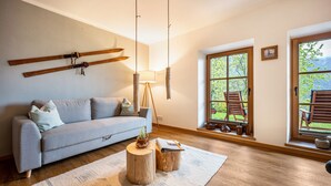 Ett vardagsrum med en soffa, träbord, en lampa och utsikt över naturen genom stora fönster.