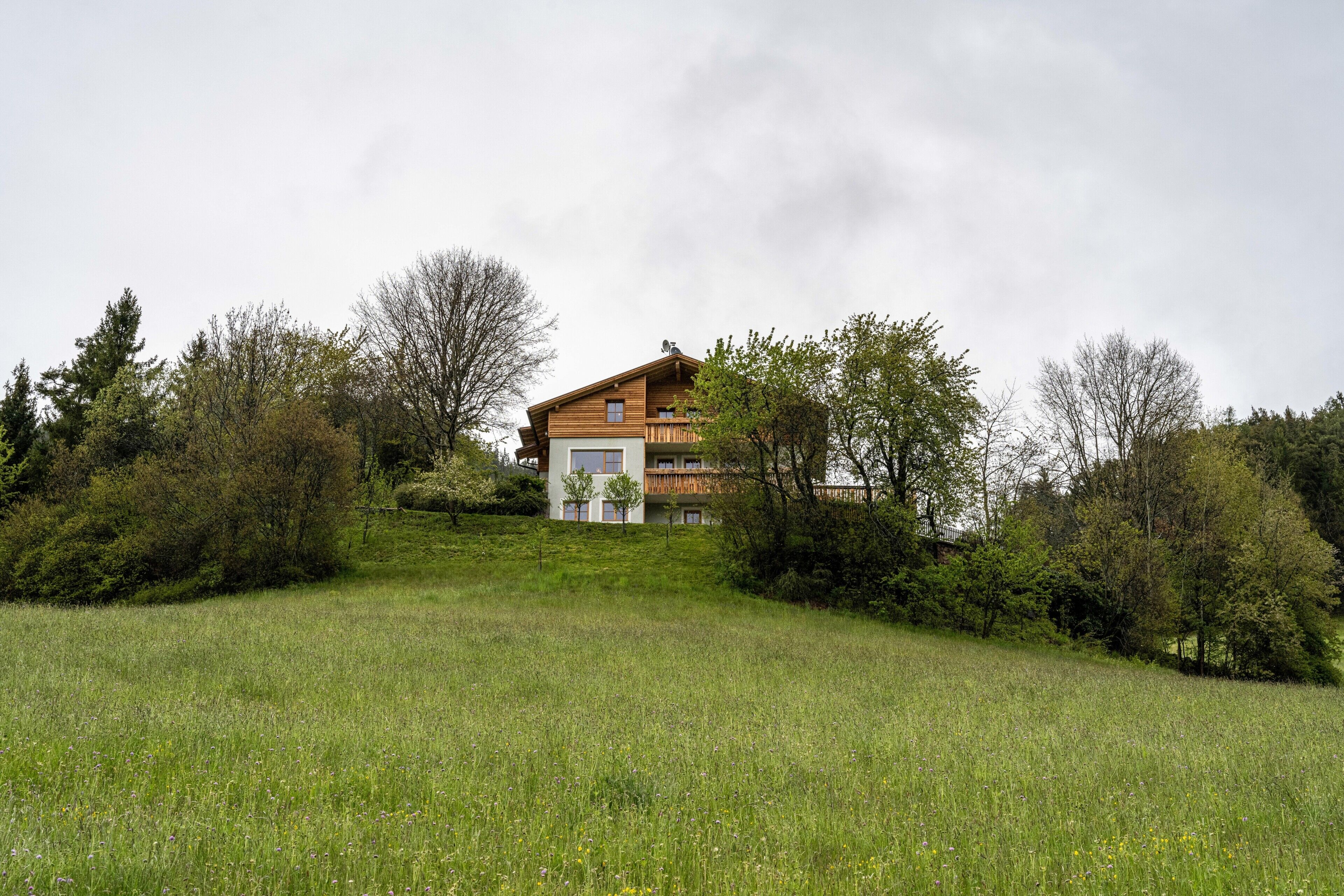 A two-story wooden house with a balcony, surrounded by a grassy field and trees.