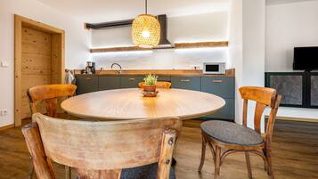 Une table ronde en bois, quatre chaises, un luminaire suspendu et un coin cuisine avec des placards et des appareils électroménagers.