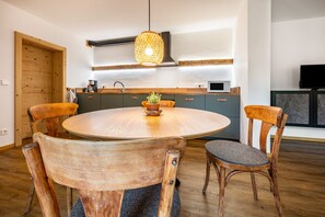 Une table ronde en bois, quatre chaises, un luminaire suspendu et un coin cuisine avec des placards et des appareils électroménagers.
