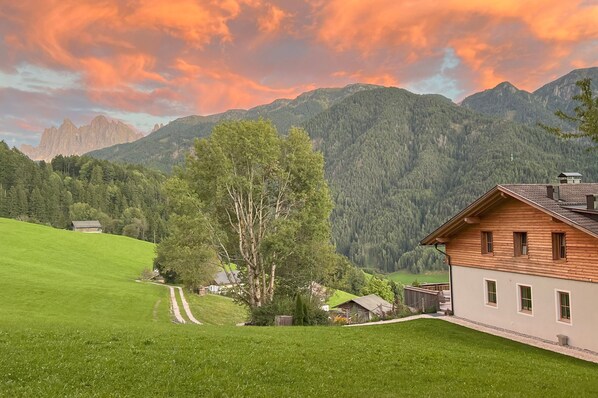 A rural house with a wooden exterior, surrounded by green fields and mountains.