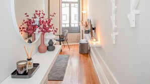 Um corredor com piso de madeira, um espelho redondo, um console branco com objetos decorativos e uma parede branca com uma obra de arte moderna.