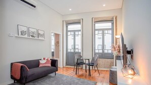 Uma sala de estar moderna com um sofá preto, tapete cinza e uma área de jantar com cadeiras e mesa. Há quadros nas paredes, uma TV em um móvel e uma janela com vista para prédios.