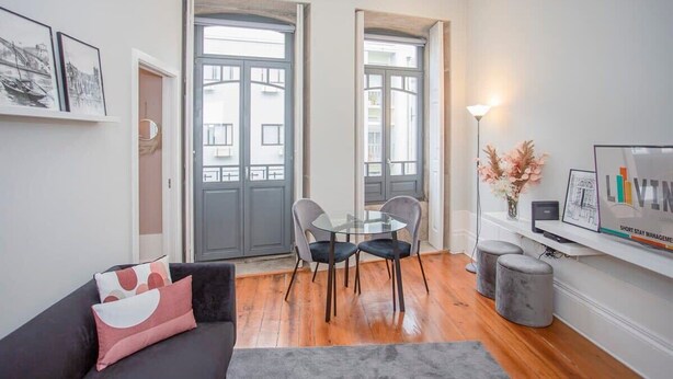 Uma sala de estar moderna com uma mesa de vidro, duas cadeiras, um sofá com almofadas decorativas, uma TV em um suporte e uma estante de livros.