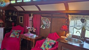 A cozy interior with a wooden ceiling, a dining table, two chairs with floral cushions, a red curtain, and a window with a heart-shaped decoration.