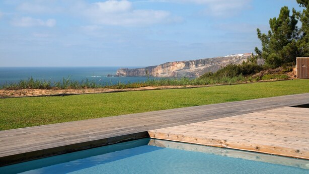 A swimming pool with a wooden deck, a grassy area, and a view of the sea and cliffs. - Apartment "Luxury Boho Style Nazaré" with Sea View, Shared Pool & Wi-Fi (Nazaré)