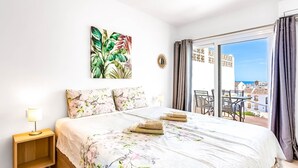 A hotel room with a bed, a nightstand, a window with curtains, a view of buildings, and a balcony with chairs. - Holiday Apartment 'Torreblanca Deluxe Holiday Home' with Sea View, Shared Pool & Wi-Fi (Fuengirola)